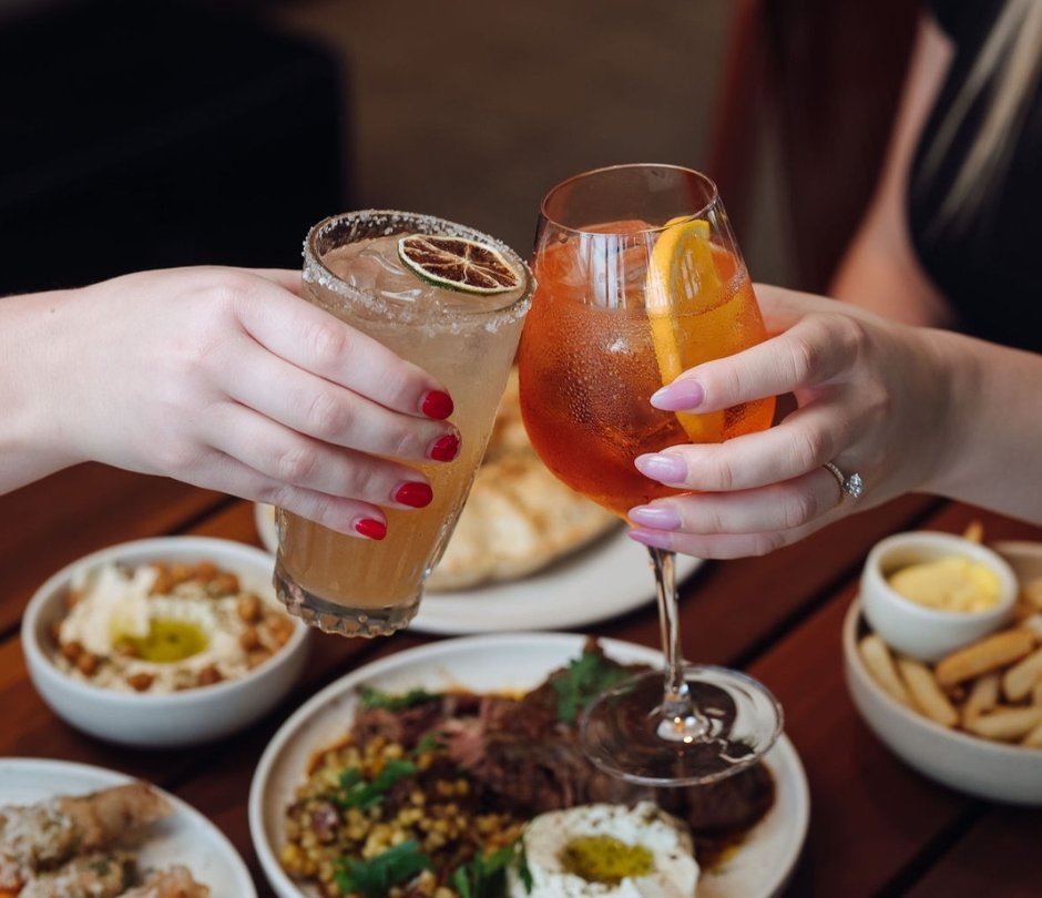 Two cocktails raised in a toast over a restaurant table filled with shared plates of food and fries.